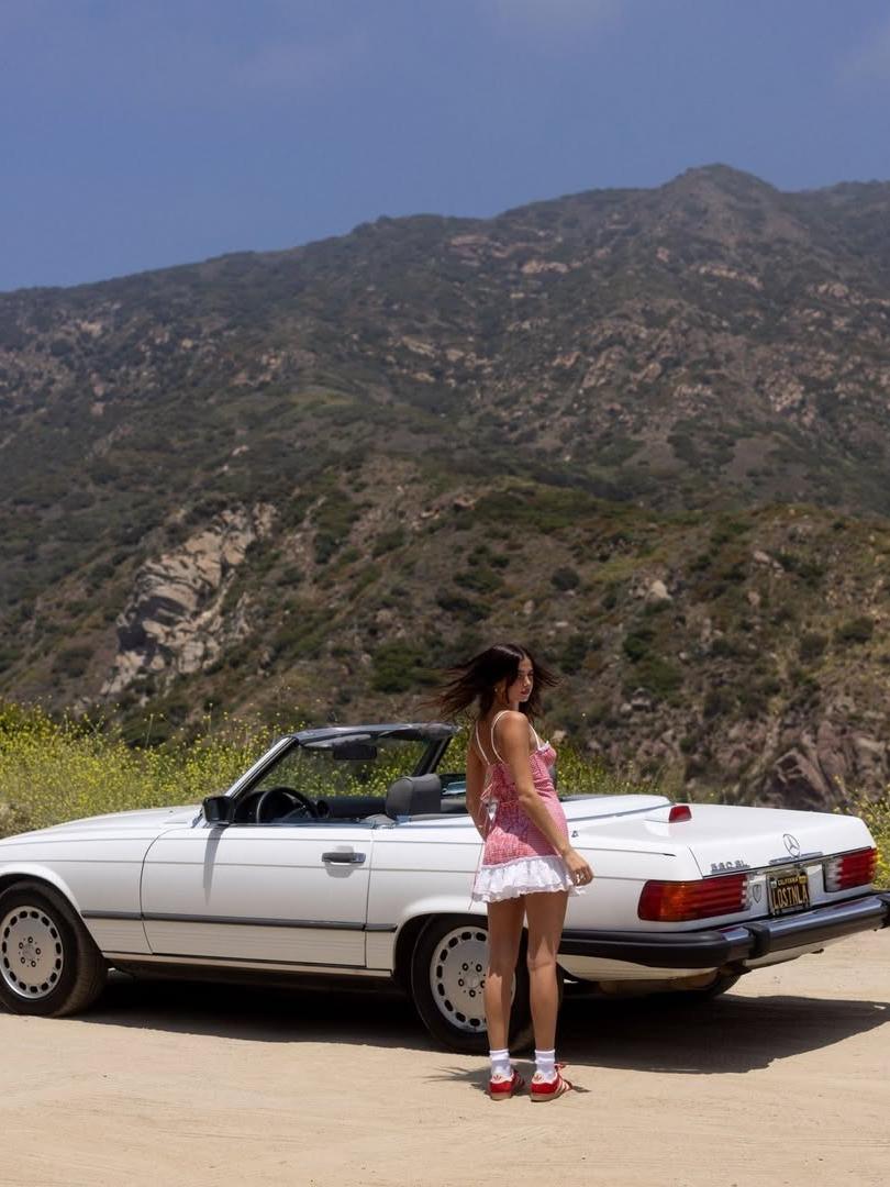 Woman in pink gingham ruffle mini dress with white trim stands beside a white convertible in a desert hillside.