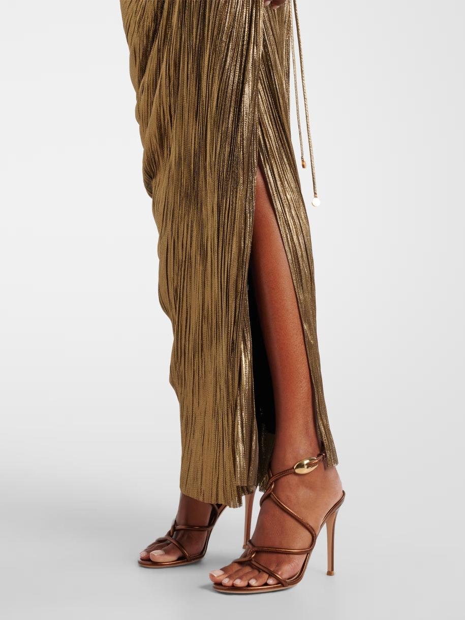 Side view of a gold metallic pleated maxi dress with a high slit, paired with brown strappy high-heeled sandals.