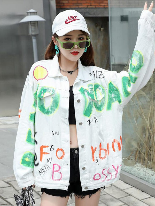 Model wearing graffiti-painted white denim bomber jacket, black crop top, white Nike cap, green sunglasses, outdoors.