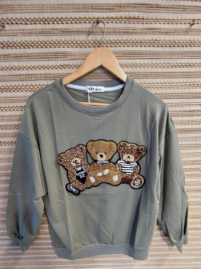 Olive green crewneck sweatshirt on wooden hanger against woven beige backdrop; sequin teddy bear graphic across chest.