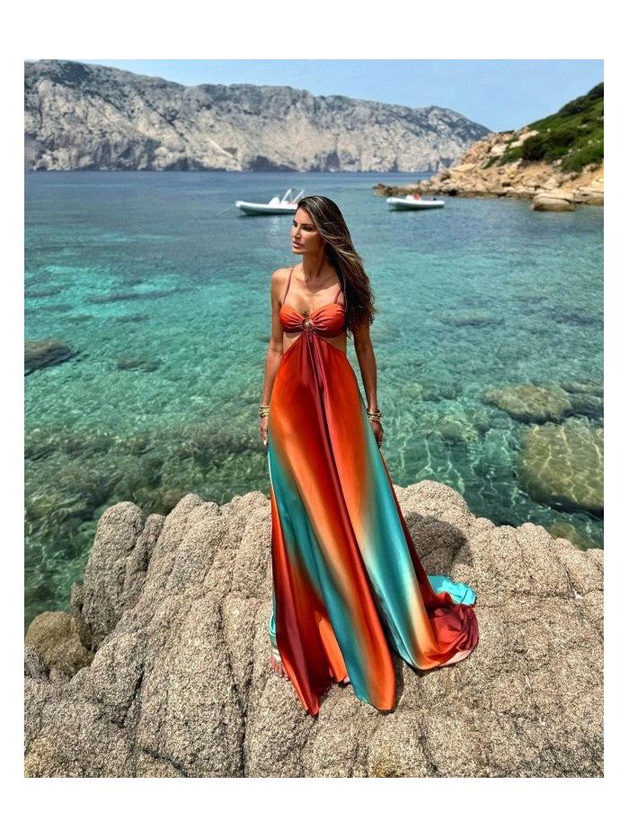 Woman in a gradient tie-dye cutout long maxi dress (red to teal) standing on rocky shore by clear turquoise sea.