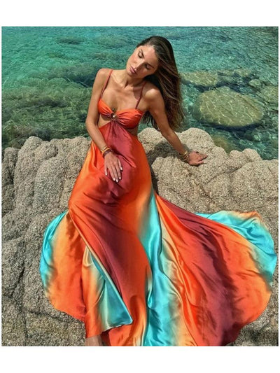 Woman wearing a gradient tie-dye cutout maxi dress with a center ring sits on a rocky shoreline beside turquoise water.