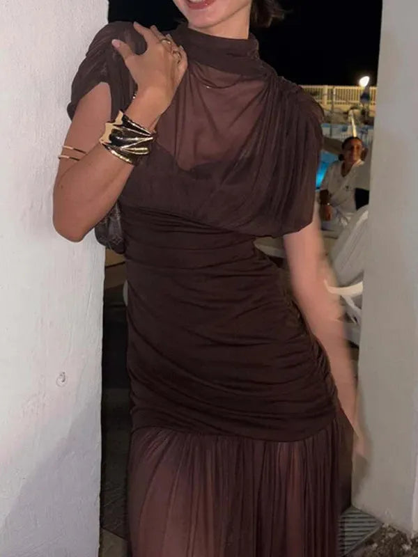 Woman in dark brown sheer mesh maxi dress with ruched bodice and pleated skirt, posing beside a white wall at night.