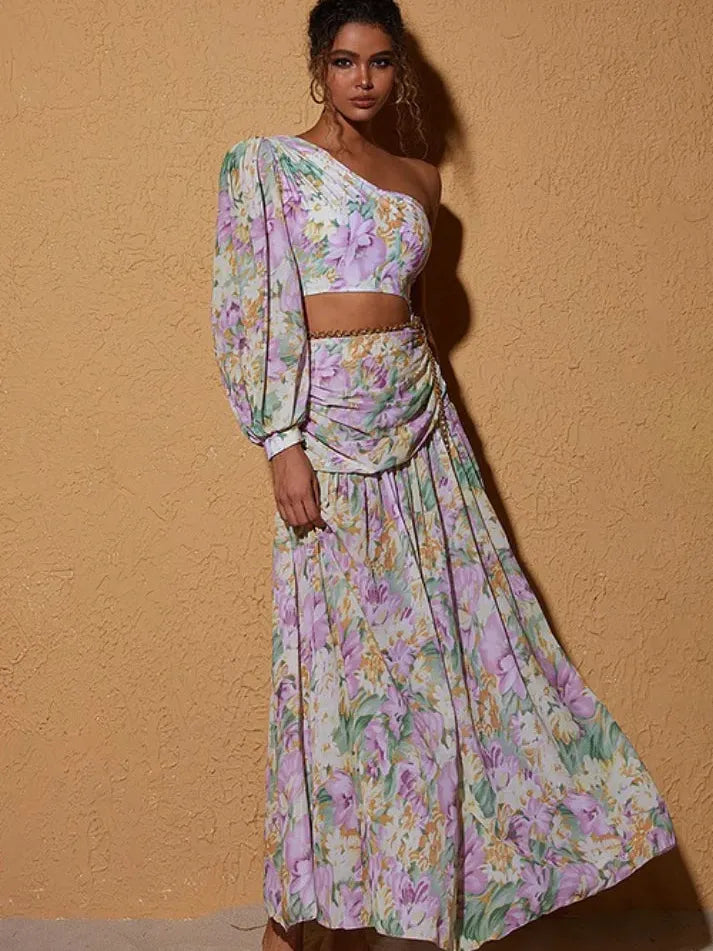 Model wearing pastel floral one-shoulder crop top with puff sleeve and matching wrap skirt; lavender and mint blossoms.