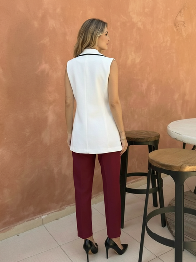 Back view of sleeveless white knit waistcoat top with black-trimmed collar, paired with burgundy pants and black heels.