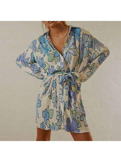 Beige pleated wrap dress with blue, green, purple floral print, long sleeves, tied at waist, pearl necklace.