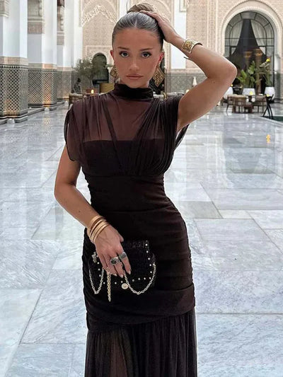 Woman wearing a dark sheer mesh ruched pleated maxi dress with high neckline and cap sleeves, in a marble courtyard.