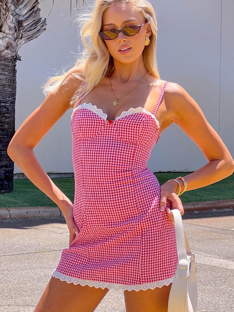 Blonde woman in red-and-white gingham mini dress with adjustable straps and white lace trim, posing with sunglasses and a purse outdoors.