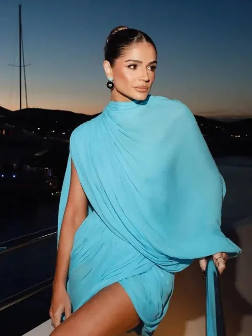 Woman in aqua draped cape mini dress with high neck, leaning on a yacht railing at sunset.