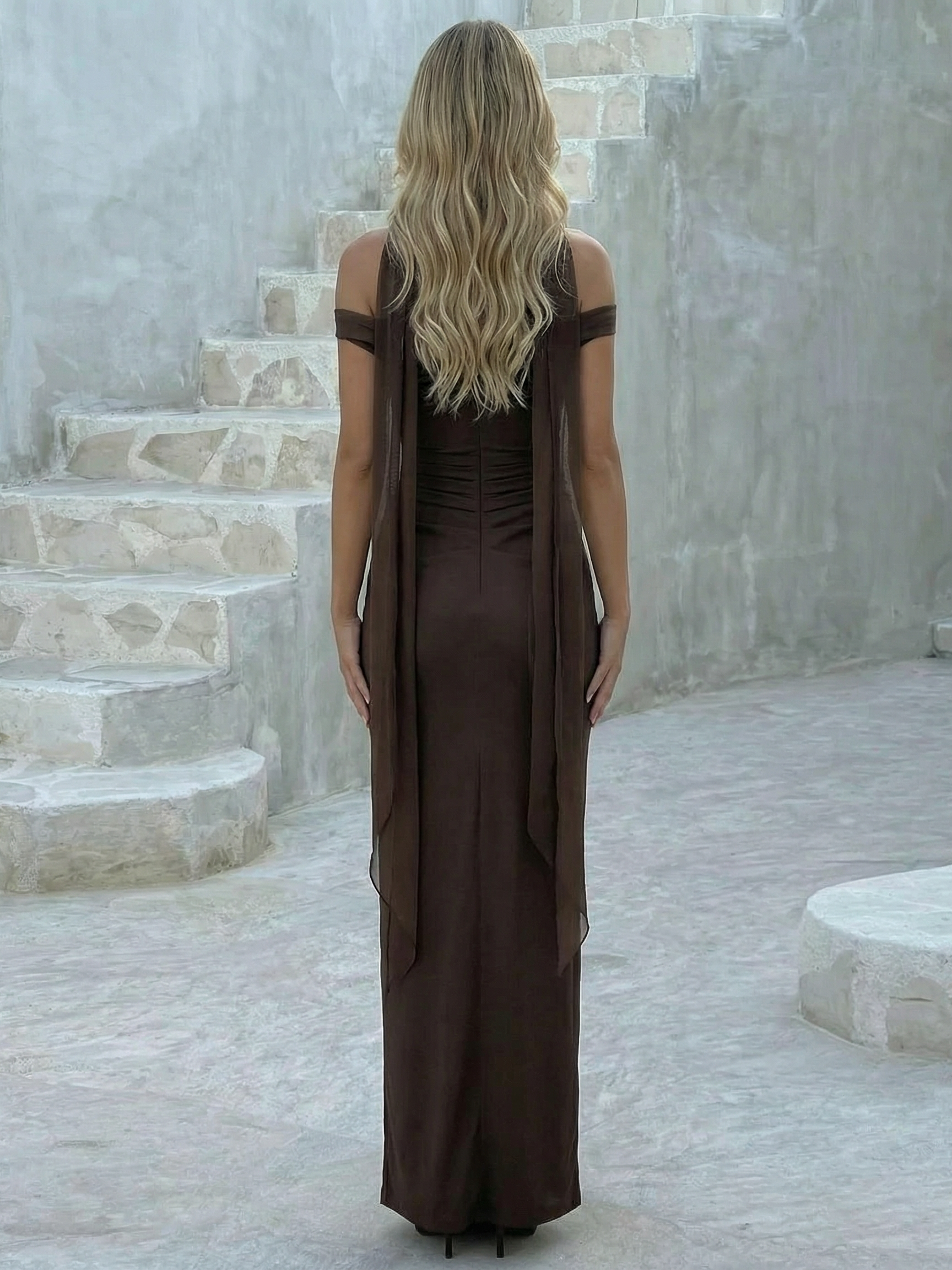 Back view of chocolate-brown off-shoulder maxi dress with long sheer scarf panels, wavy blonde hair, on stone stairs.