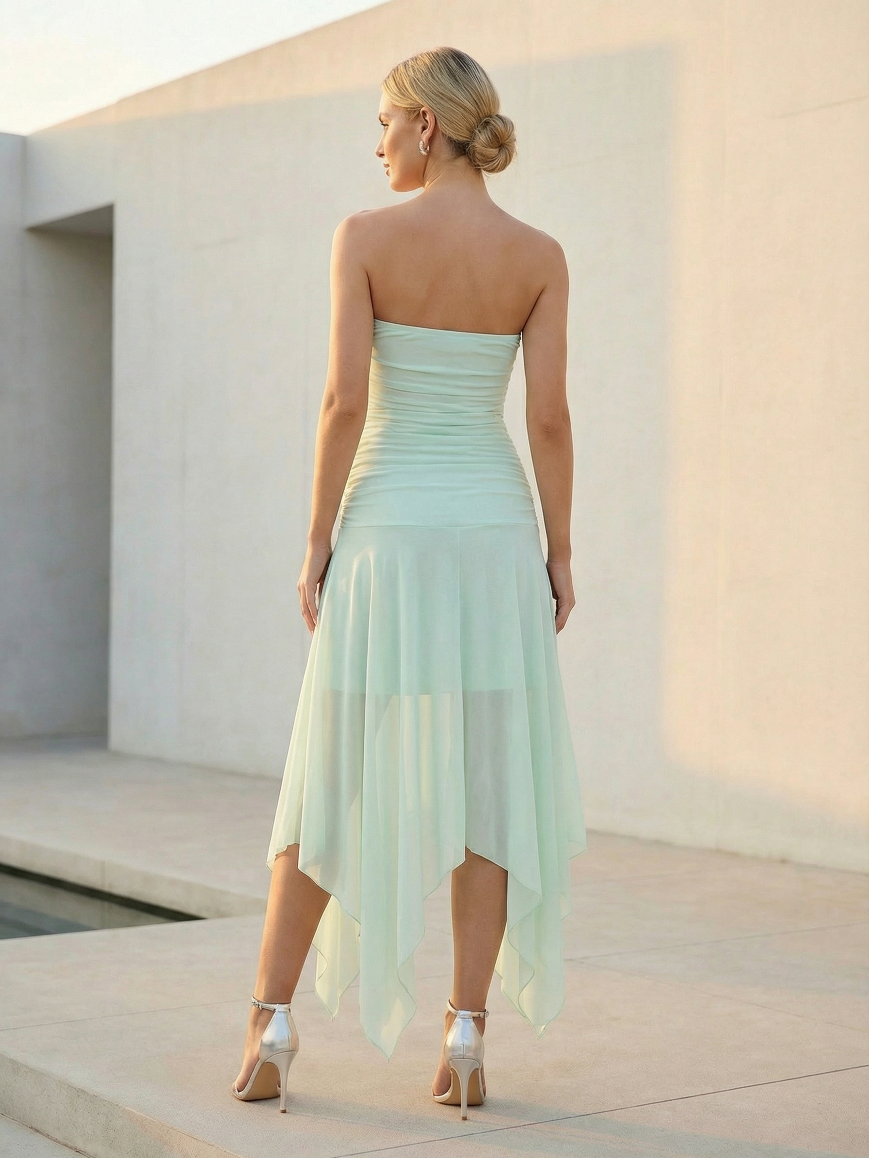 Back view of a woman wearing a mint green strapless ruched chiffon dress with a handkerchief hem, and silver heels.