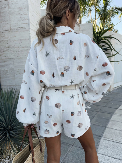 Back view of white seashell-print shirt with oversized sleeves and matching belted shorts; model with woven tote.