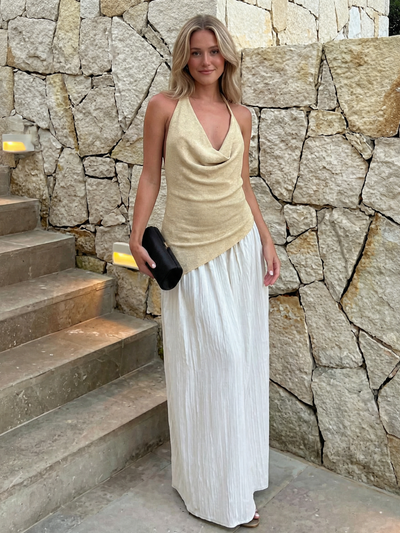 Blonde woman in beige draped halter top with white pleated maxi skirt, holding a black clutch beside a stone wall.