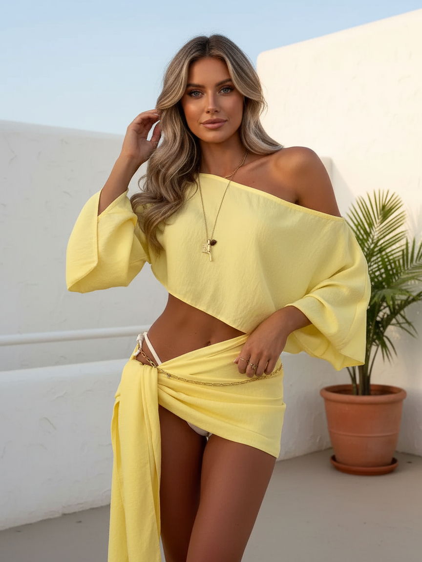 Woman in yellow off-shoulder draped bikini cover-up with asymmetrical hem and matching wrap skirt with gold chain belt.