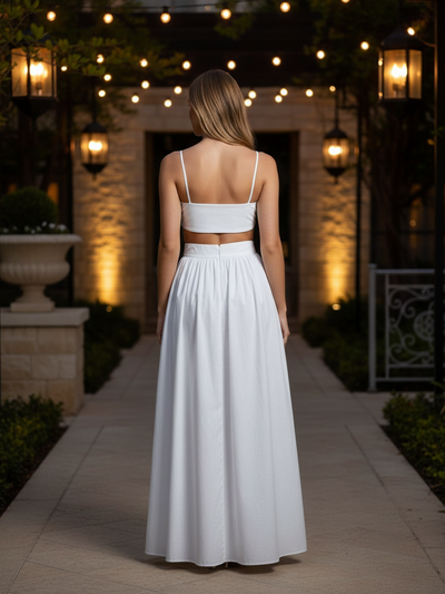 Back view of a woman in white spaghetti-strap crop top and high-waisted maxi skirt on a stone path under string lights.