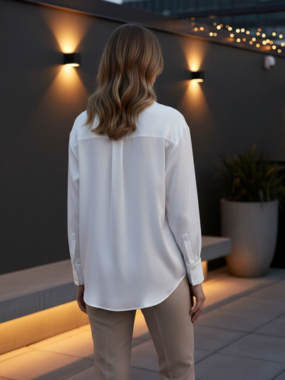 Back view of a white satin-feel button-down shirt with long sleeves and curved hem, paired with beige trousers outdoors.