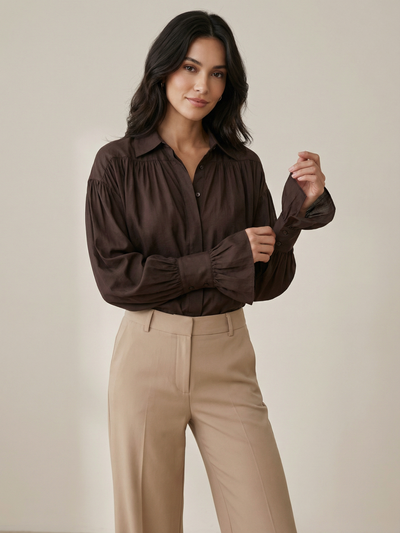 Woman wearing a brown oversized shirt with puff sleeves, gathered yoke and ruffled cuffs, paired with beige trousers.