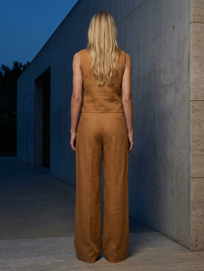Back view of a woman in camel linen vest top and waistcoat with wide-leg trousers, outdoors beside a concrete wall.