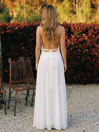 Back view of a blonde woman in a white halter-neck draped maxi dress with waist tie, standing on gravel beside a rusted bench.
