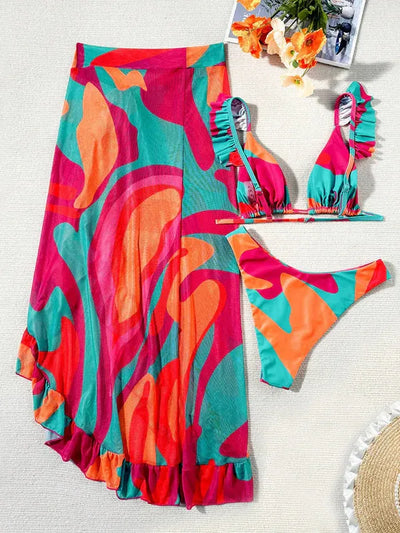 Bright abstract-print bikini with ruffled teal-pink top, matching bottom, and a wrap skirt on a beige background.