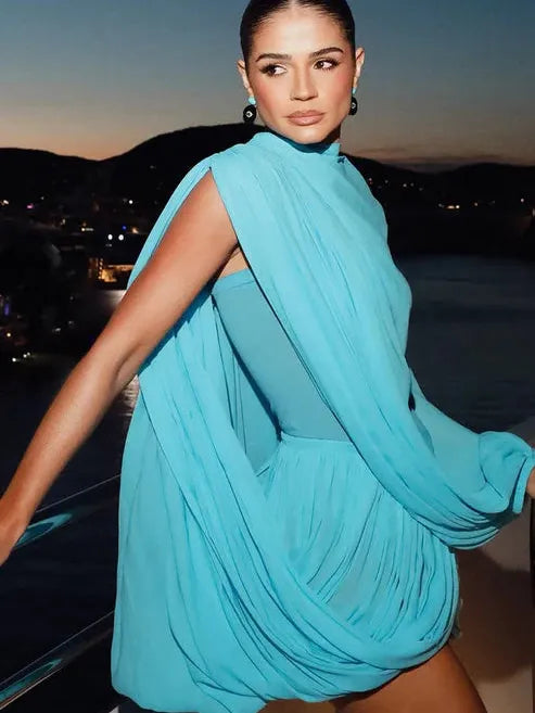 Turquoise draped cape mini dress on a model; flowing cape panels over a fitted bodice, by water at dusk.