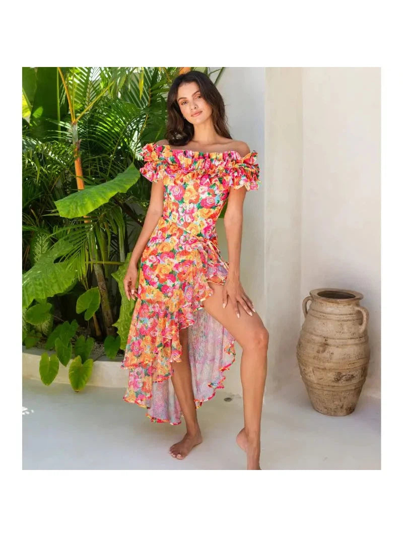 Model wearing a colorful floral off-shoulder ruffle bodysuit with a high-low skirt swimsuit, barefoot beside plants.