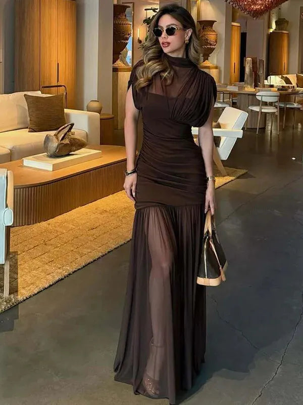 Woman in a dark brown sheer mesh ruched pleated maxi dress with a high neckline and sheer overlay, posed in chic lounge.