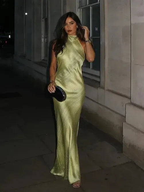 Woman in lime-green striped halter-neck maxi dress with a knotted waist, walking at night and holding a black clutch.