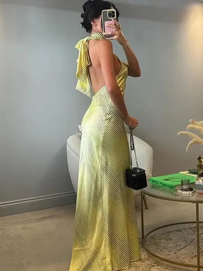 Side view of a woman in a yellow striped halterneck maxi dress with an open back and knotted back tie, carrying a black purse.