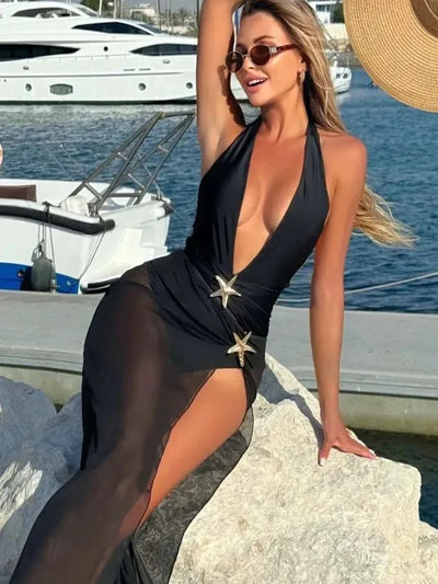 Black halter-neck swimsuit with sheer sarong skirt and starfish belt, model sits on a rock by the marina.