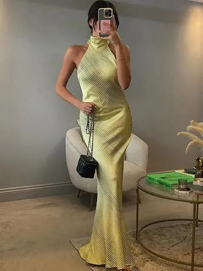 Woman in lime-yellow striped halter-neck maxi dress with a side knot, indoors, carrying a black quilted handbag.