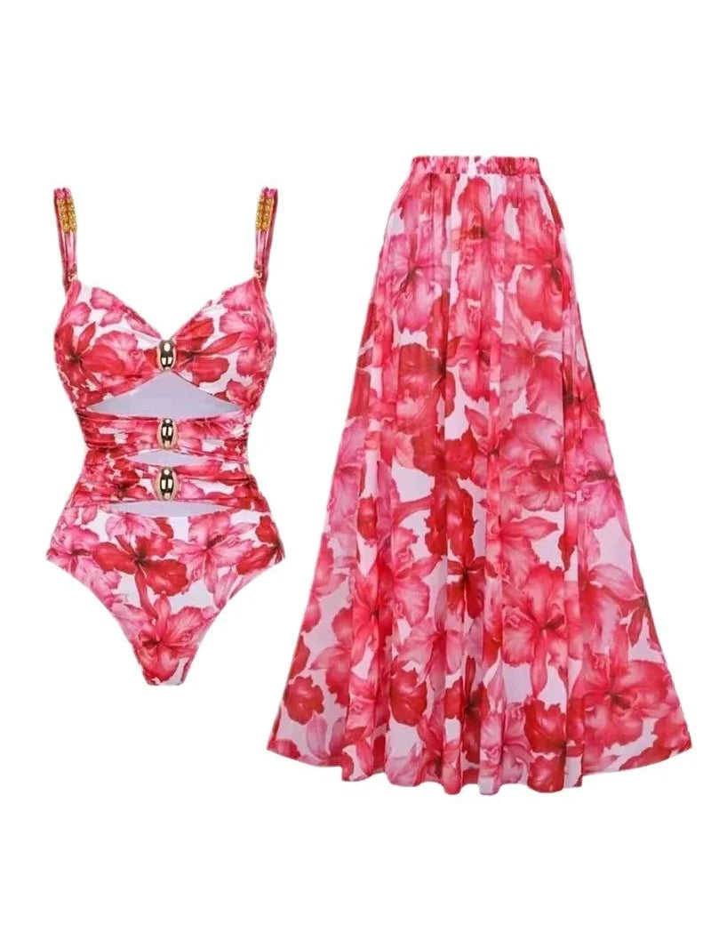 Floral pink one-piece swimsuit with cutout midsection and gold buckle accents on straps, paired with sheer floral skirt.