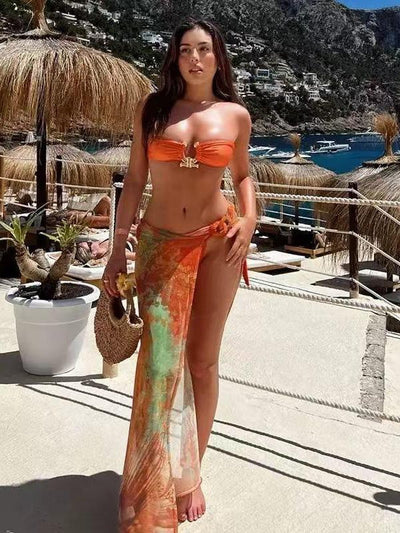 Sunlit beach: woman in orange bandeau bikini and tie-dye sarong, barefoot, holding a straw bag, with thatched umbrellas and blue sea behind.