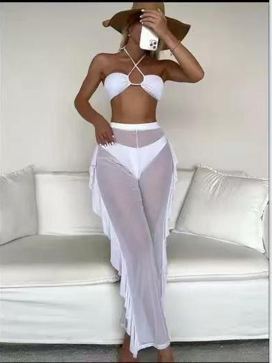 Model wearing white criss-cross halter bikini top with ring, matching bottoms, and sheer white cover-up pants; brown wide-brim hat.