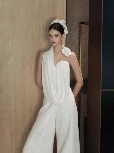 Model in white draped one-shoulder co-ord with wide-leg pants, floral shoulder accent, and white hair bun accessory.