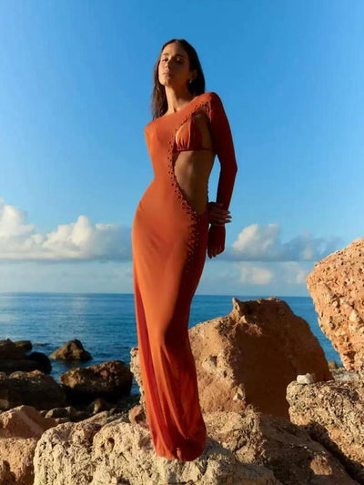 Model in burnt-orange one-shoulder halter bikini and long sarong with side cutout and beaded trim, on rocky shore.