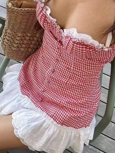 Red gingham ruffle mini dress with white lace trim, thin straps, worn on a chair with a woven straw bag on a wooden deck.