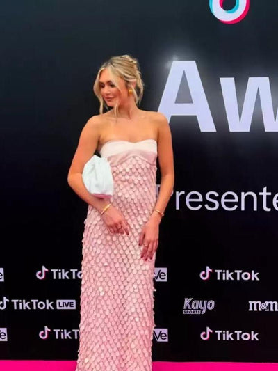 Blonde on pink carpet in a strapless pink mermaid-sequin maxi dress with scale-like sequins, holding a blue clutch.