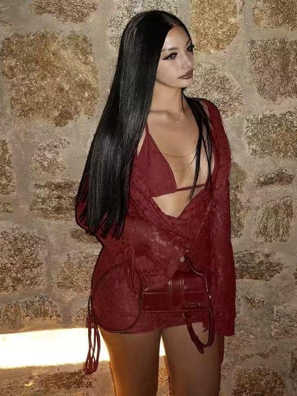 Woman in burgundy lace cut-out halter mini dress with drawstring sleeves, long black hair, carrying a small burgundy crossbody bag against a stone wall.
