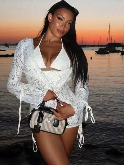 Woman in white lace cut-out halter mini dress with long drawstring sleeves, holding a woven handbag at sunset by the water.