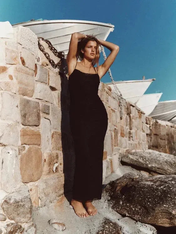 Model in a black spaghetti-strap ruched midi dress, barefoot, by stone wall on a rocky beach with boats in the background.