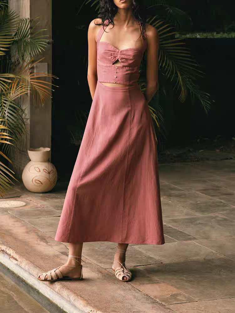 Woman in pink halter-neck cut-out maxi dress with button bodice and A-line skirt, beige sandals, garden backdrop.