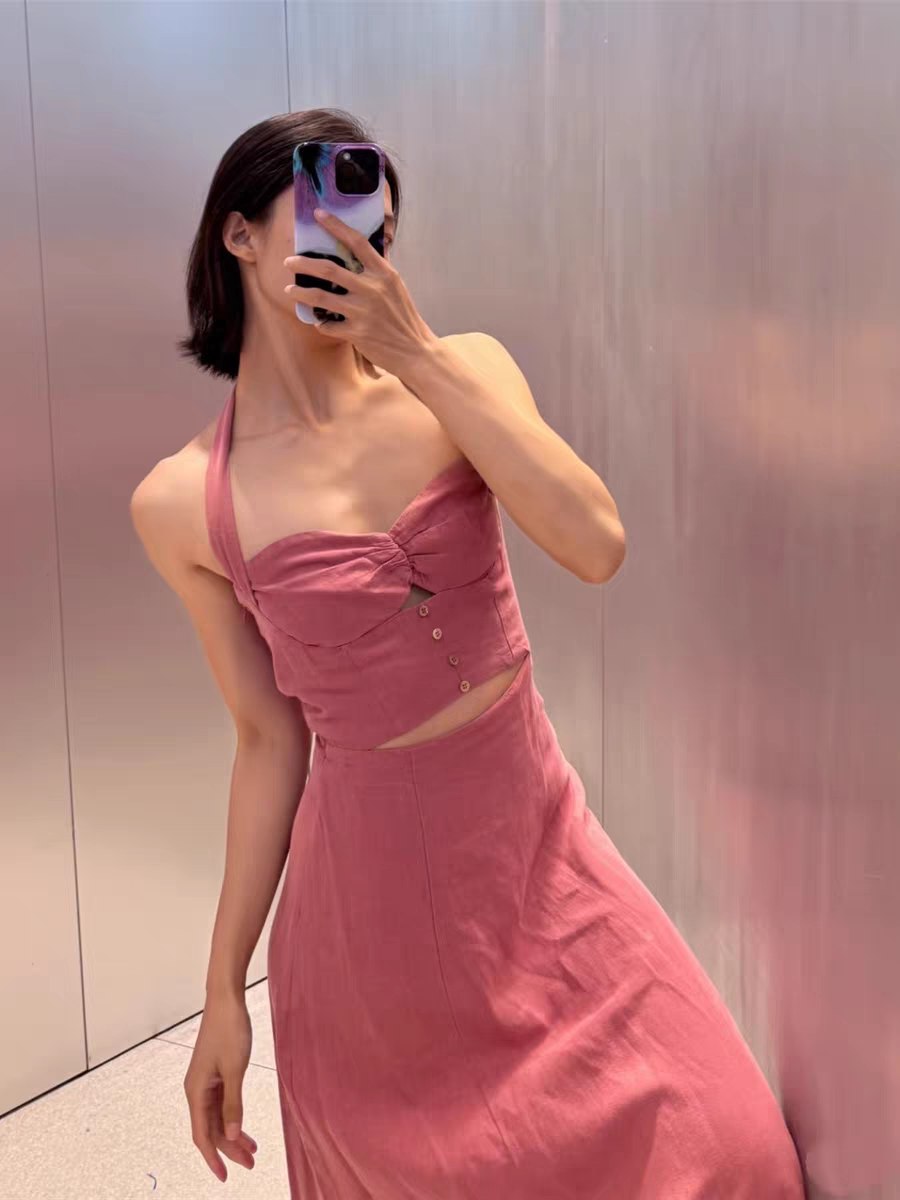 Woman in pink halter-neck cut-out maxi dress, midriff-baring with front buttons, posing for a mirror selfie against a metallic wall.