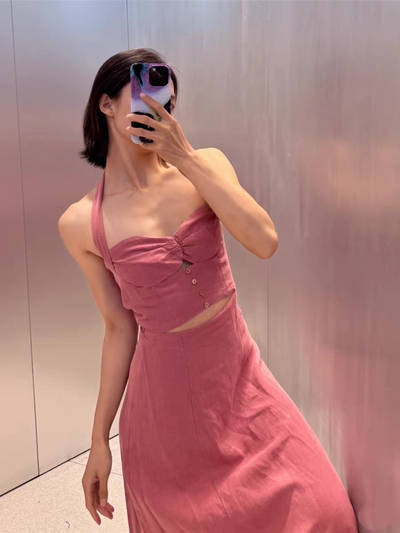 Woman in pink halter-neck cut-out maxi dress, midriff-baring with front buttons, posing for a mirror selfie against a metallic wall.
