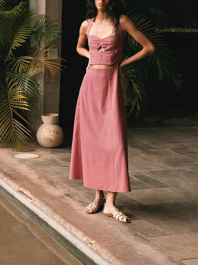 Pink halter-neck cut-out maxi dress with front buttons, worn by a woman standing on a stone patio among tropical plants.