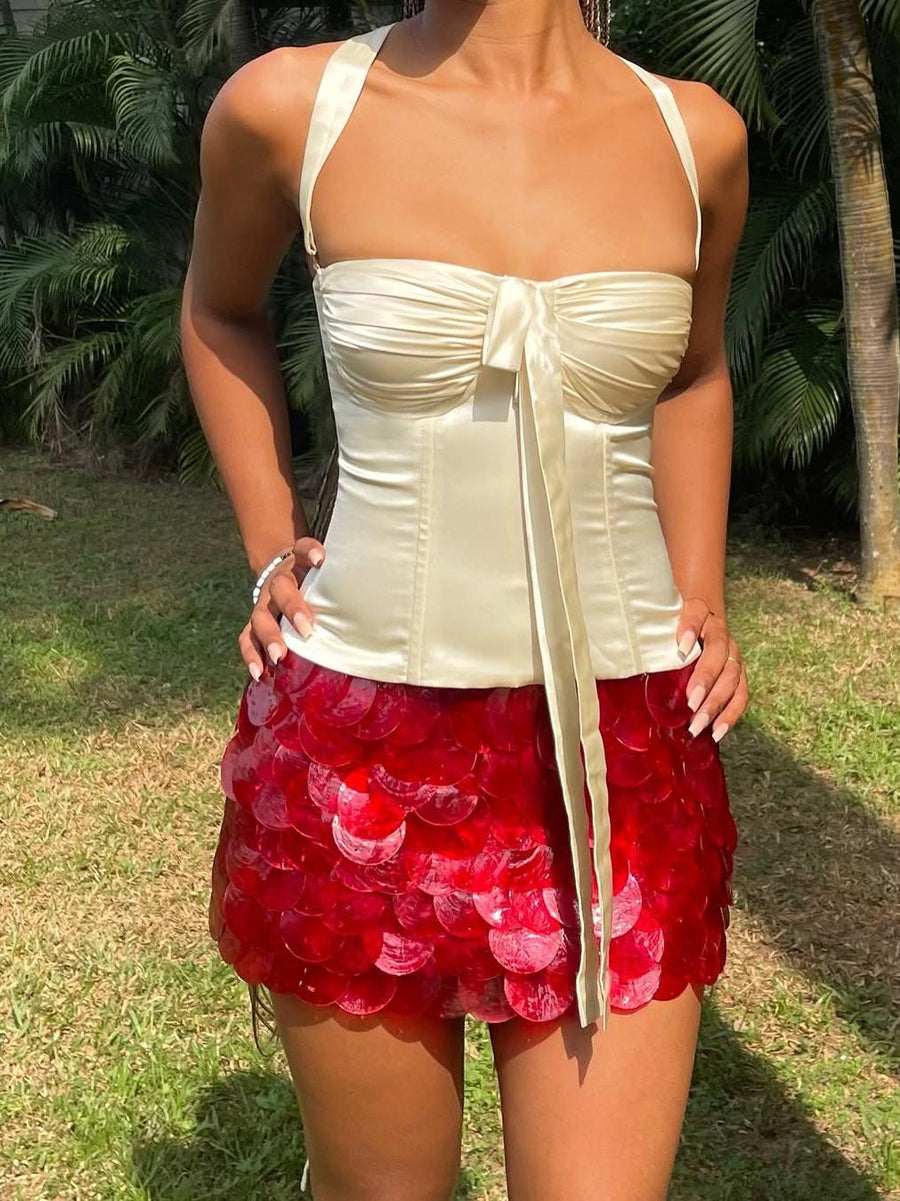 Cream satin halter top with front bow paired with a red coin-shell mini skirt of overlapping sequins, outdoors.
