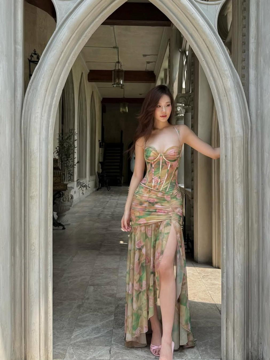 Model in pastel floral tie-dye corset dress with ruched bodice and sheer high-slit skirt, pink heels in a stone archway.