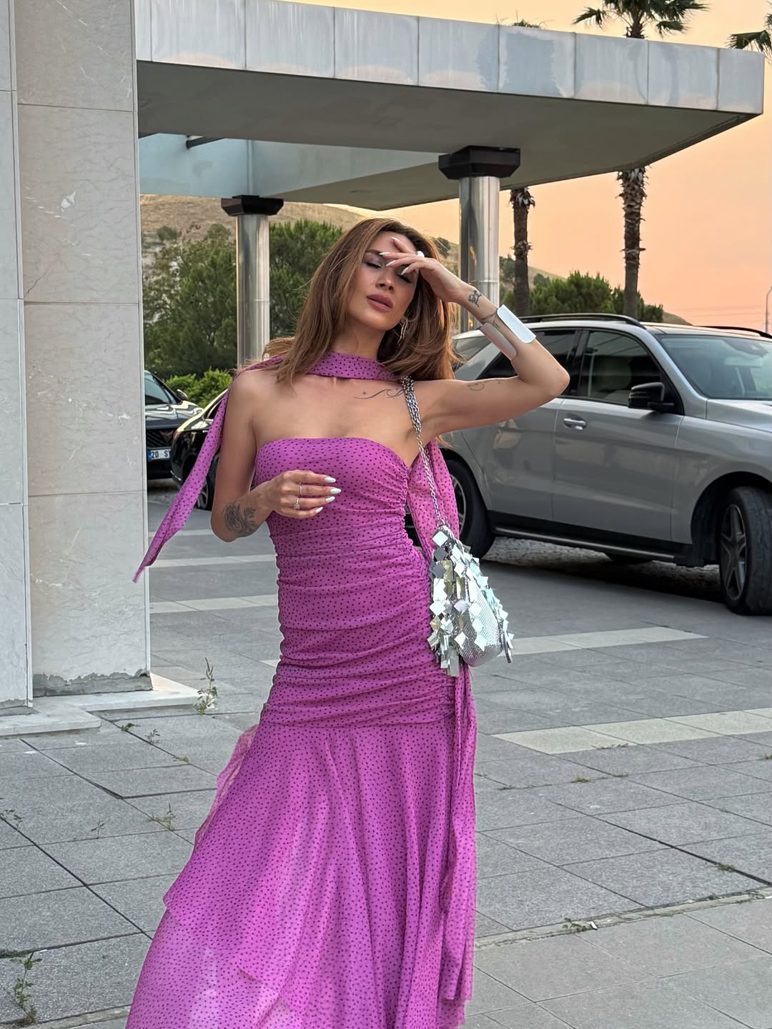 Magenta polka-dot, one-shoulder ruched dress with asymmetric scarf draped at the neck, tiered ruffle mermaid hem, silver mirrored handbag.