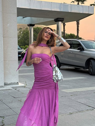 Magenta polka-dot, one-shoulder ruched dress with asymmetric scarf draped at the neck, tiered ruffle mermaid hem, silver mirrored handbag.