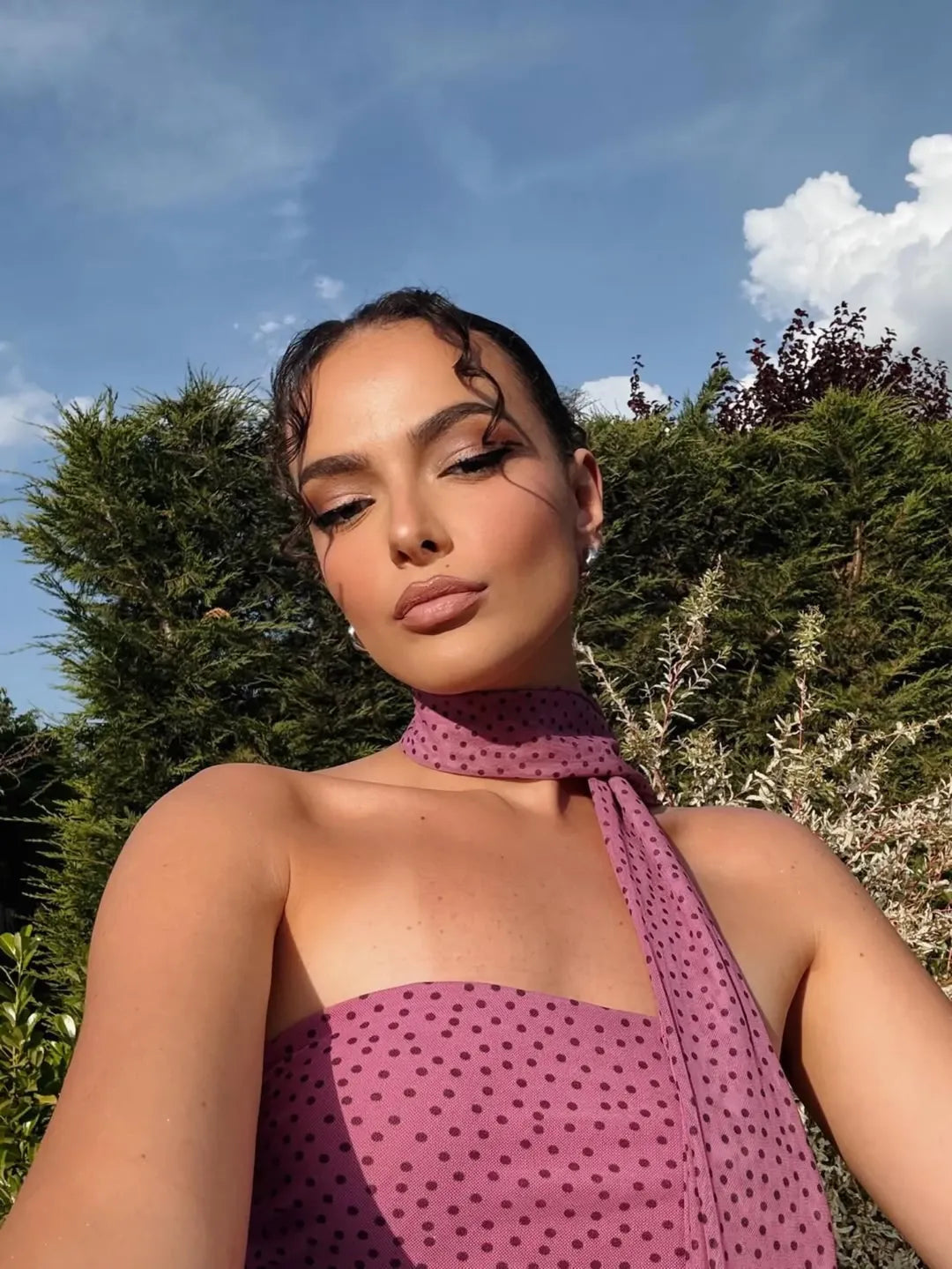 Portrait of a woman in a pink polka-dot strapless dress with a scarf tied at the neck, outdoors among greenery.